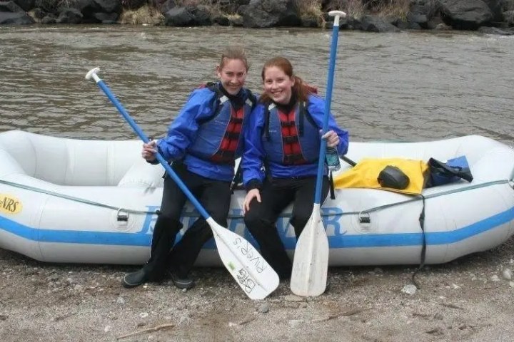 Two people in blue jackets stand by an inflatable raft near a river, holding paddles.