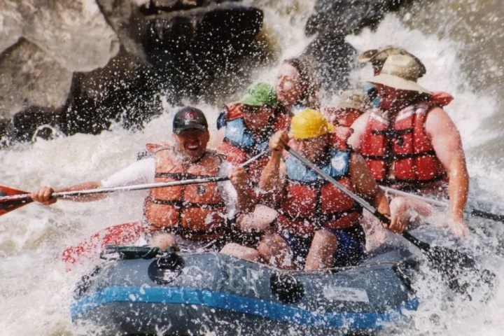 Group of people whitewater rafting in an inflatable boat, wearing helmets and life vests, splashed by waves.