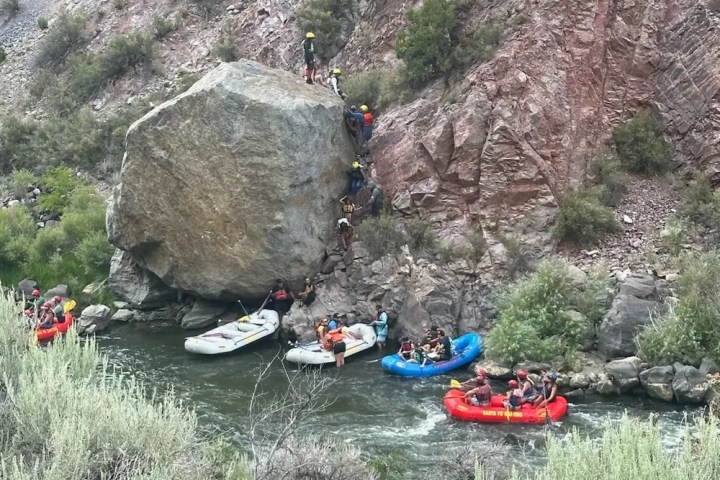People rafting on a river beside a rocky cliff with vegetation.