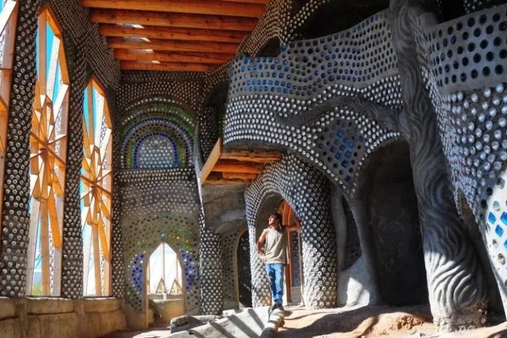 Person standing inside a colorful building with bottle walls and wooden ceiling.