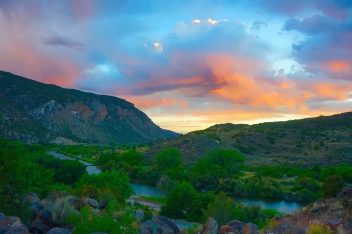 River valley with green trees, mountains, and colorful sunset sky.